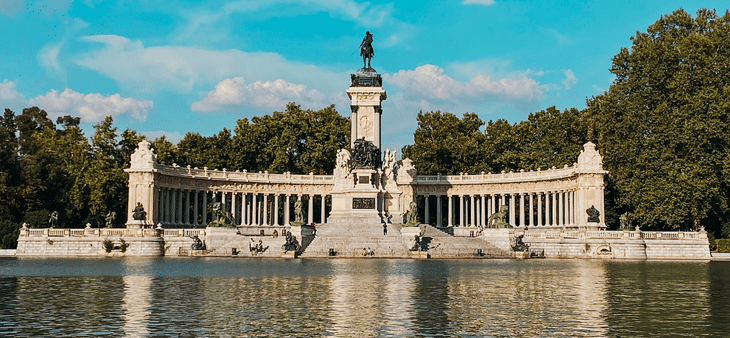 Monumento a Alfonso XII reflejado en el Estanque Grande del Retiro de Madrid, rodeado de árboles y cielo azul.