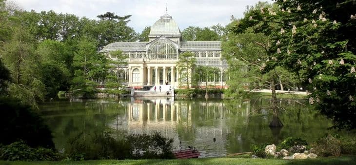 Palacio de Cristal del Parque del Retiro de Madrid reflejado en el estanque, rodeado de árboles y vegetación.