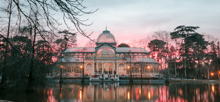 Palacio de Cristal del Parque del Retiro de Madrid al atardecer, reflejado en el estanque entre árboles y cielo rosado.