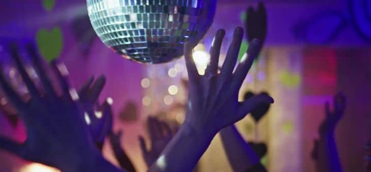 Manos levantadas bailando bajo una bola de espejos durante una fiesta nocturna con luces de colores.